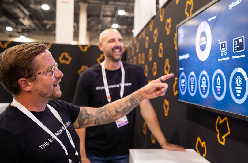 A Splunker pointing at a monitor with graphics on it, as another Splunker looks on.