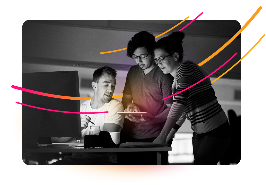 Three people are collaborating around a computer monitor in an office setting. One person is seated, while two others stand behind, looking at the screen and a tablet.