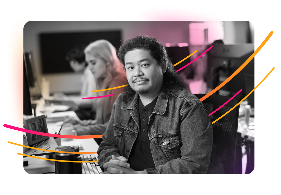 A man with long hair and a beard, wearing a denim jacket, sits at a desk and looks at the camera, with other people working in the background and abstract pink and orange lines overlaid on the image.